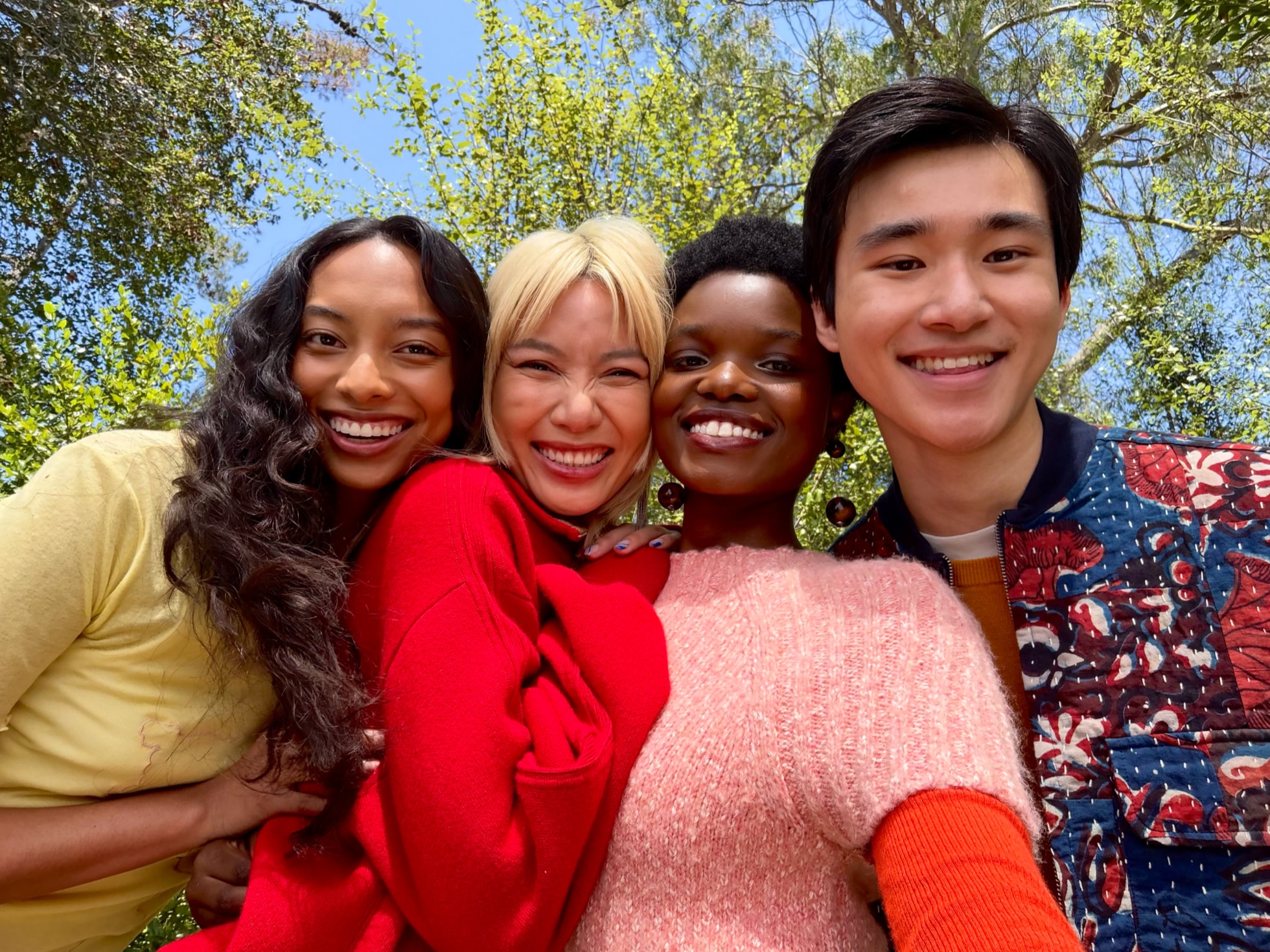 Four smiling friends fitting into one wide group selfie under green trees and blue sky